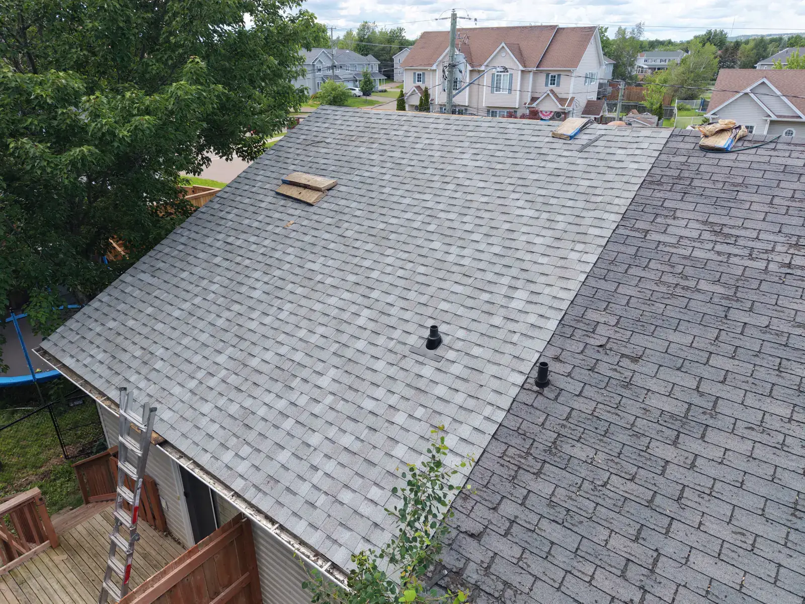 Mid-install at this Moncton duplex: new shingle courses down one face, bundles staged at the ridge, ladder set at the eave