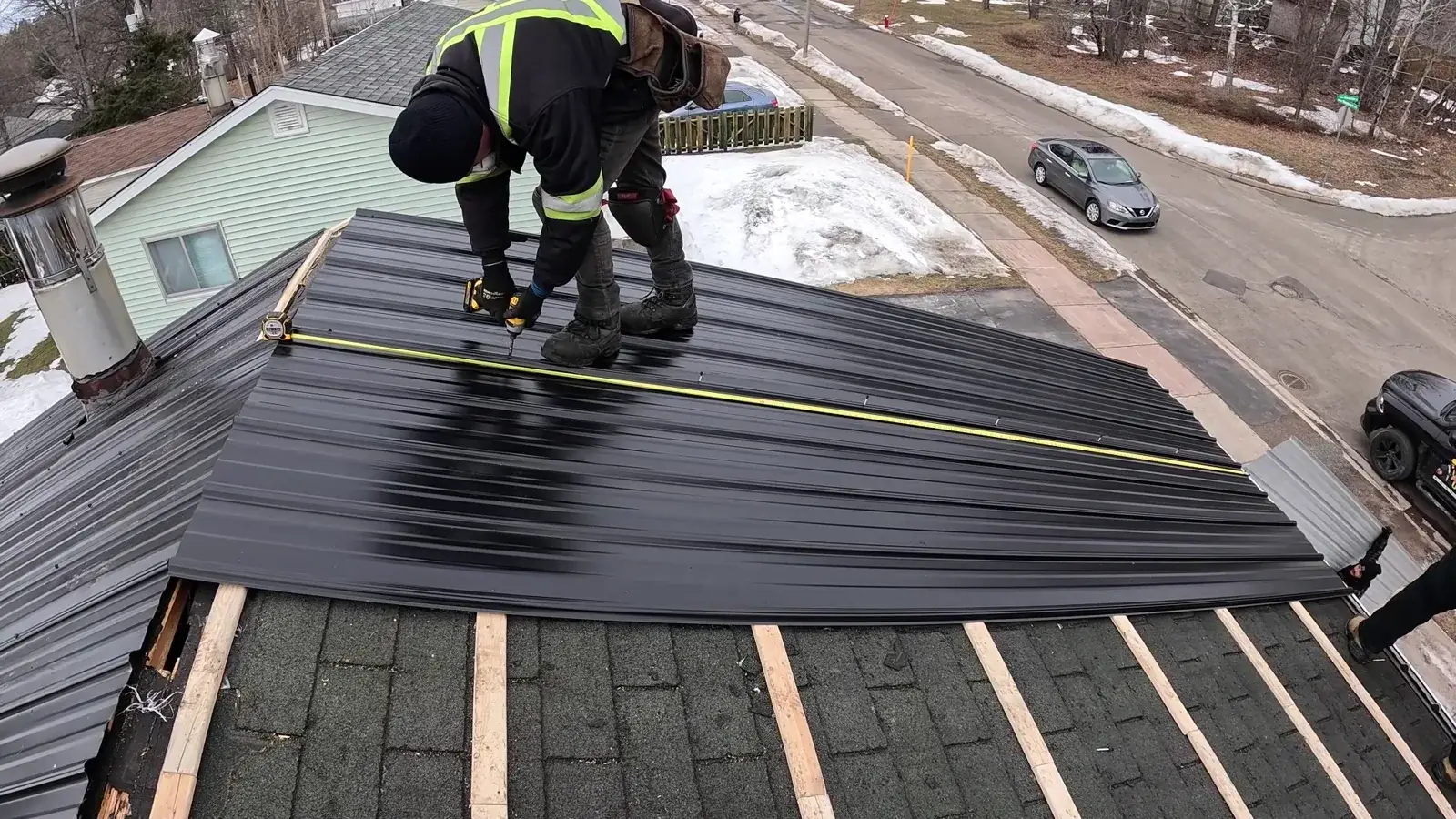 Roofer fastening charcoal ribbed steel panels over strapping during a winter metal install in New Brunswick